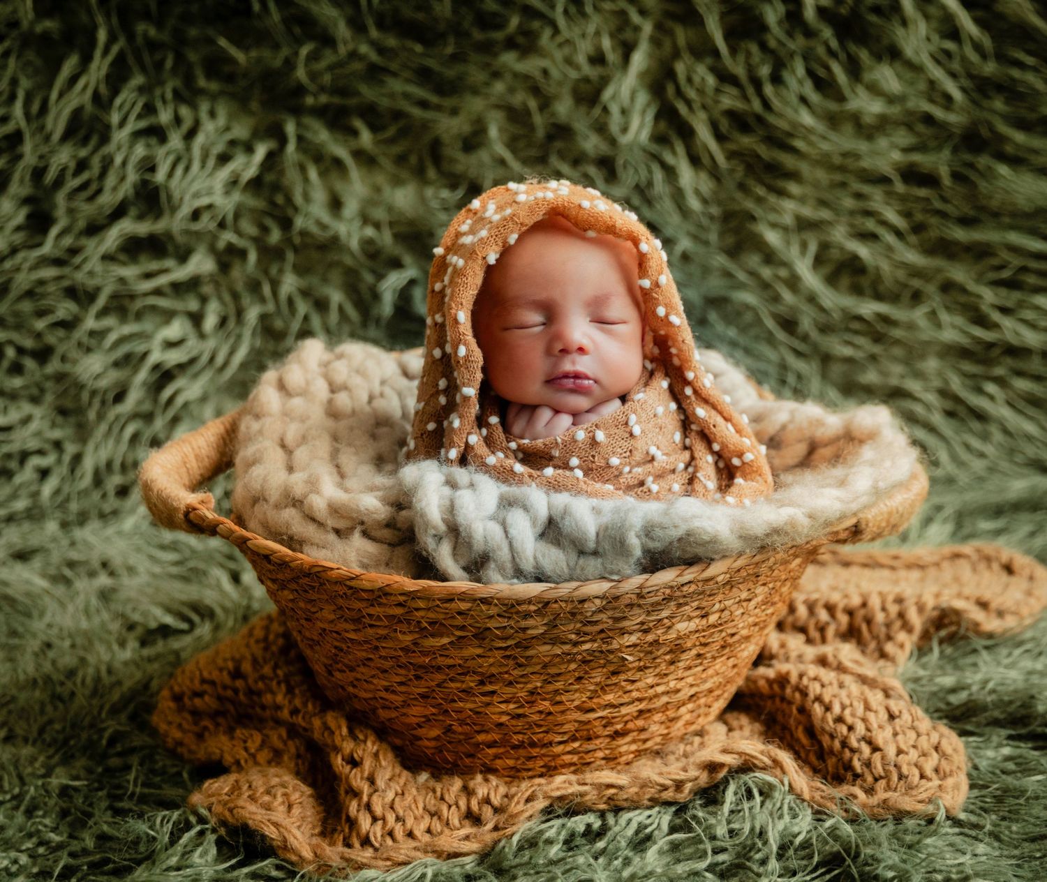 Newborn baby sleeping peacefully in a wicker basket with a brown polka dot wrap on a green fur background.