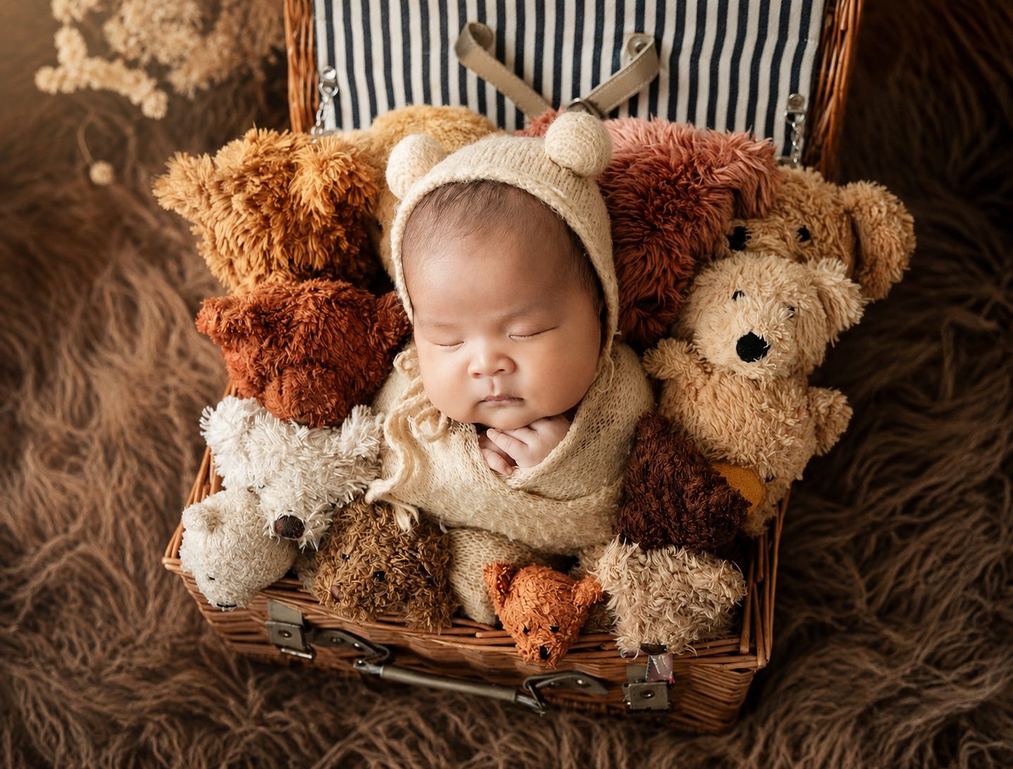 Sleeping newborn baby in bear bonnet nestled among teddy bears in a wicker basket on fur backdrop.