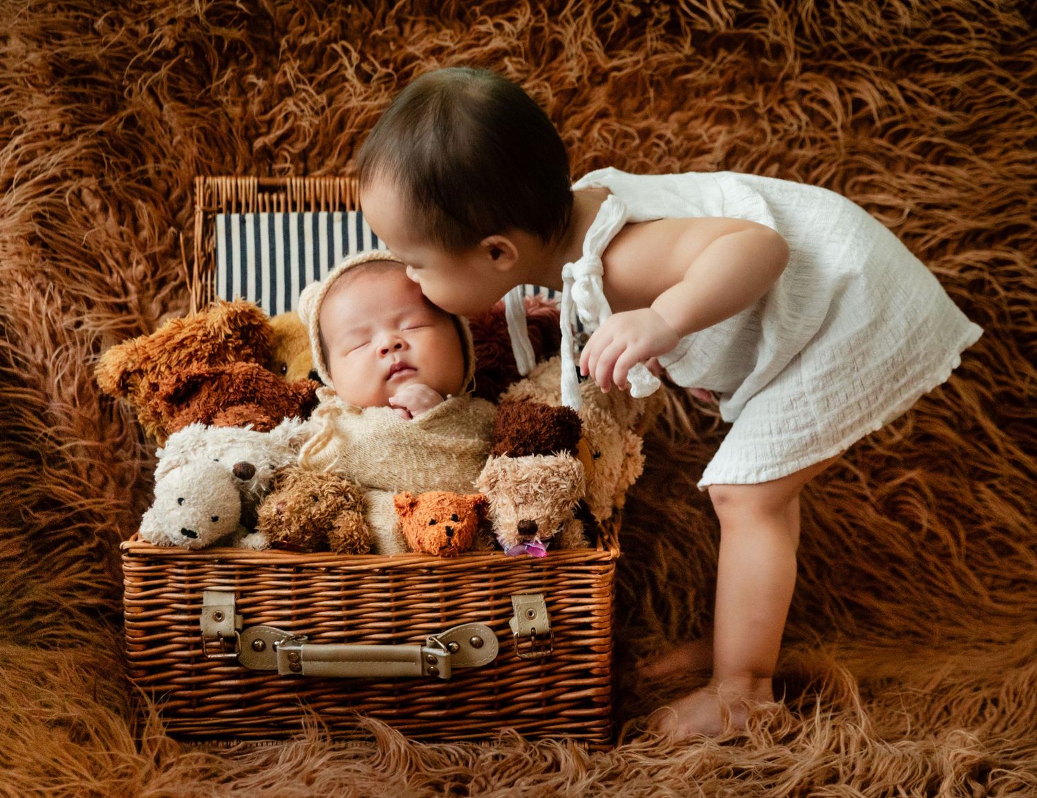Toddler girl in white dress kissing newborn baby sibling nestled in wicker suitcase surrounded by teddy bears.
