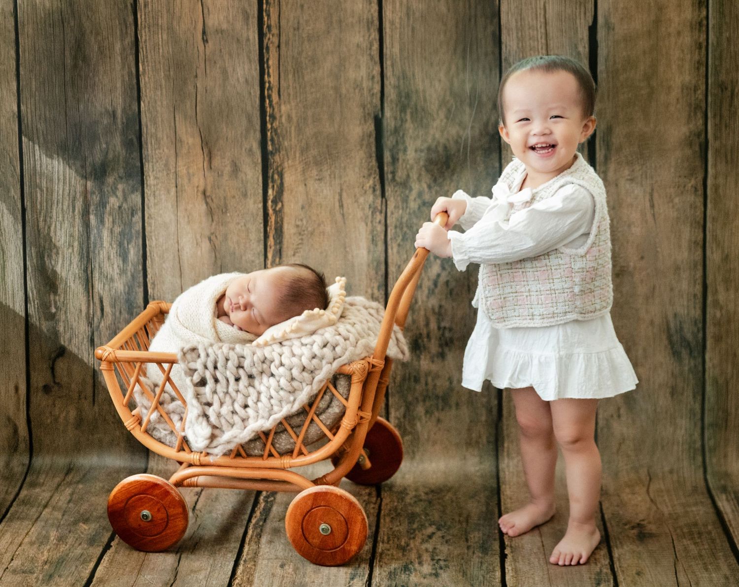 Toddler girl smiling while pushing a wicker baby carriage with a sleeping newborn inside, against a rustic wood backdrop.