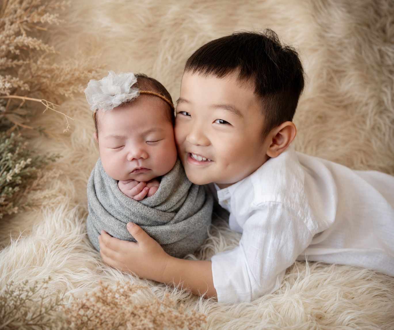 Young boy smiling while gently holding a sleeping newborn baby wrapped in a gray swaddle on a fluffy beige fur rug.