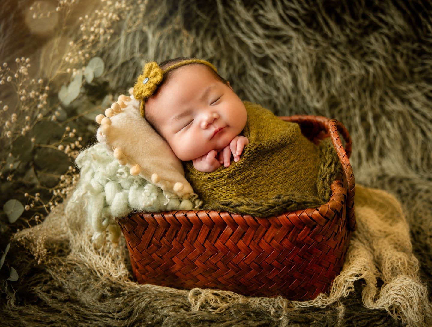 Smiling newborn baby wrapped in green swaddle sleeping peacefully in a rustic red wicker basket with floral decor.