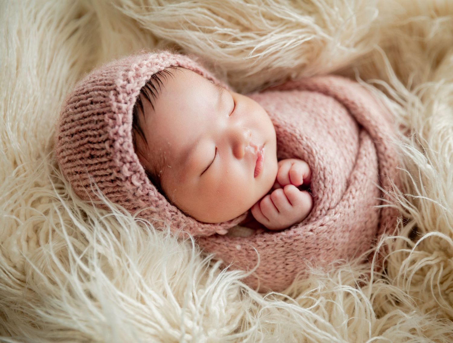 Sleeping newborn baby wrapped in a soft pink knit blanket and bonnet, nestled on a fluffy cream fur rug.