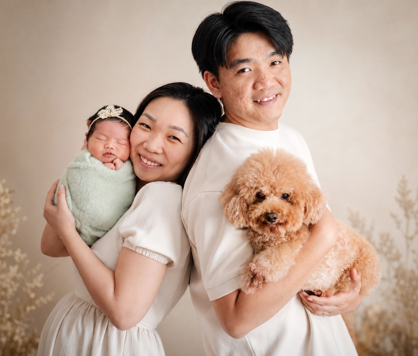 A smiling couple poses for a newborn photo shoot, holding a swaddled baby and a fluffy golden poodle.