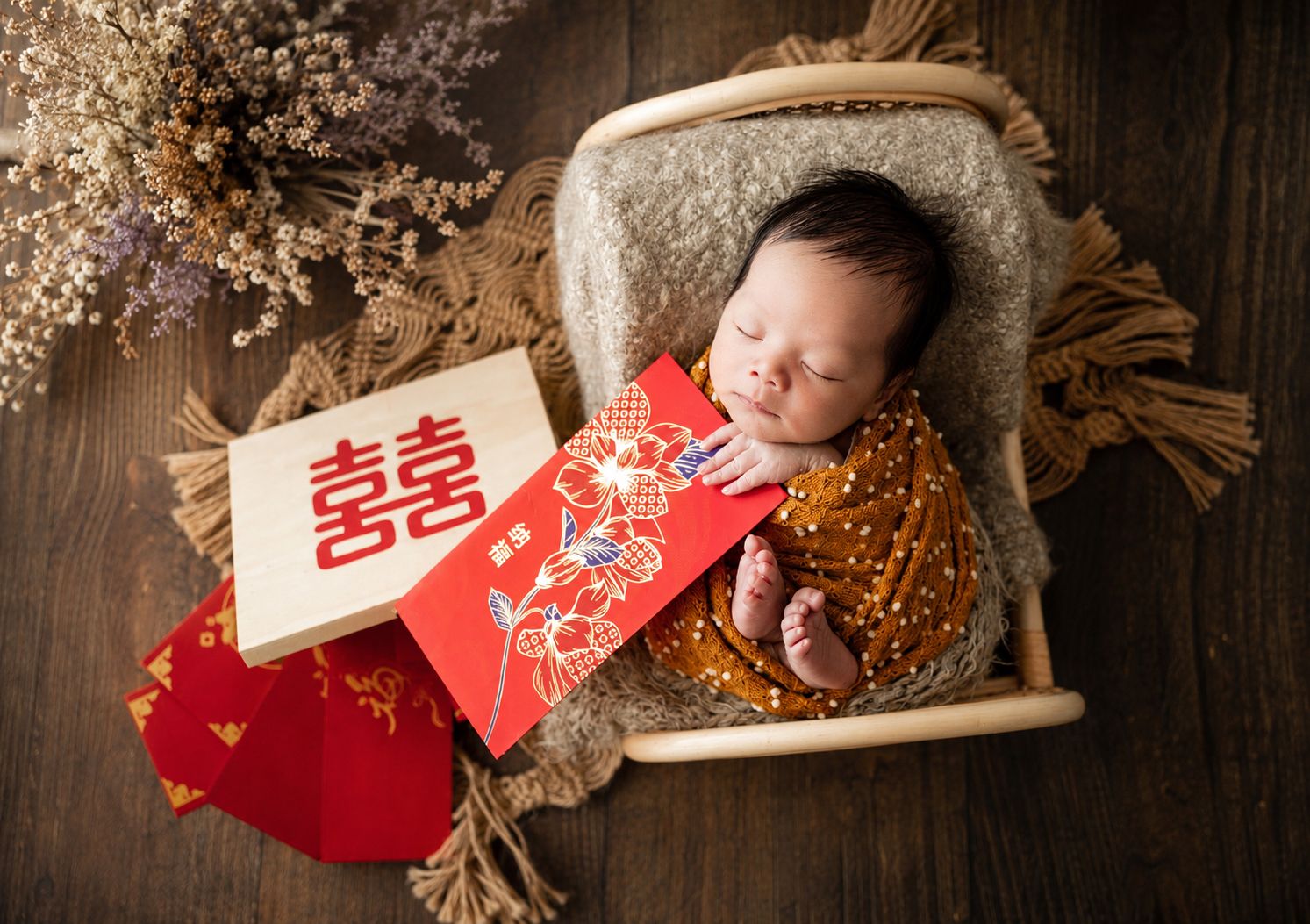 Newborn baby wrapped in gold swaddle sleeping in wicker basket with Chinese red envelopes and double happiness symbol.