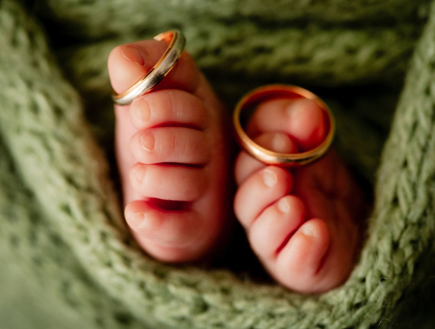 Newborn baby hands holding two gold wedding rings wrapped in green knit blanket.