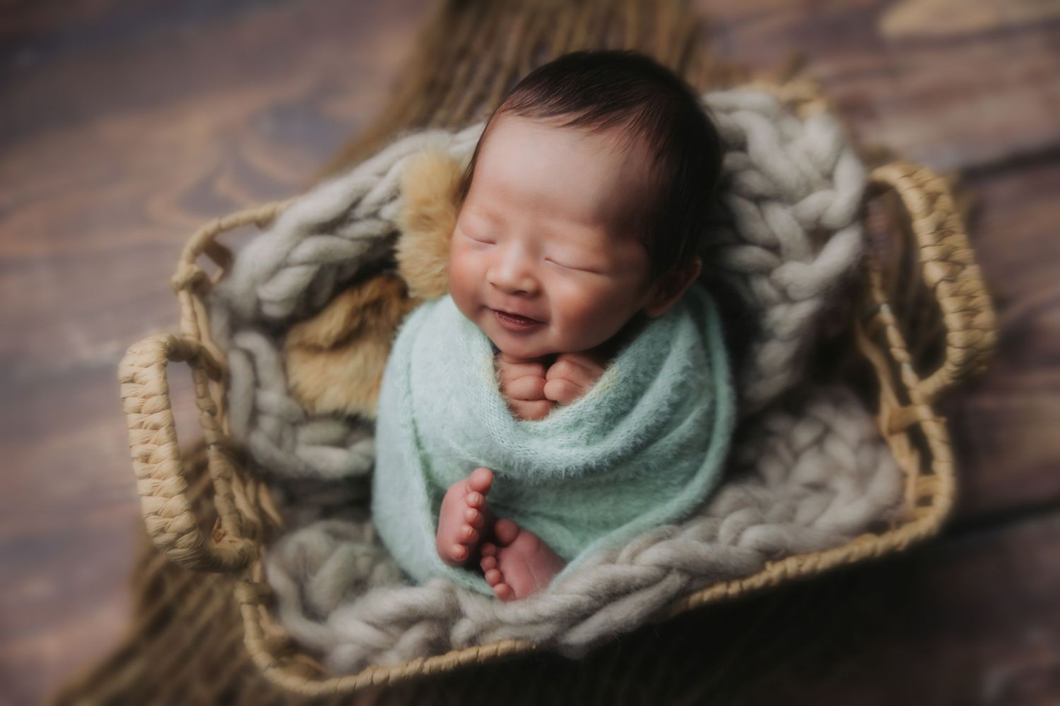 Smiling newborn baby wrapped in mint green swaddle sleeping in a wicker basket with chunky knit blanket.