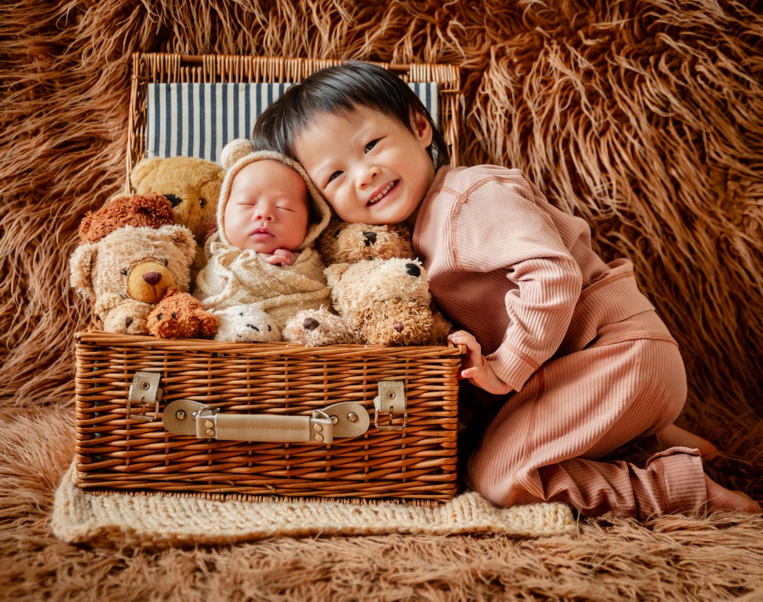 Smiling toddler poses with sleeping newborn baby nestled in wicker basket surrounded by teddy bears on fluffy brown fur.