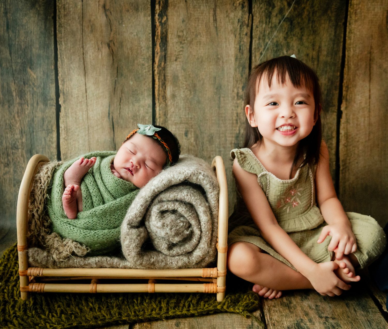 Smiling toddler girl in green dress sits beside a sleeping newborn wrapped in green swaddle in a small wooden bed.