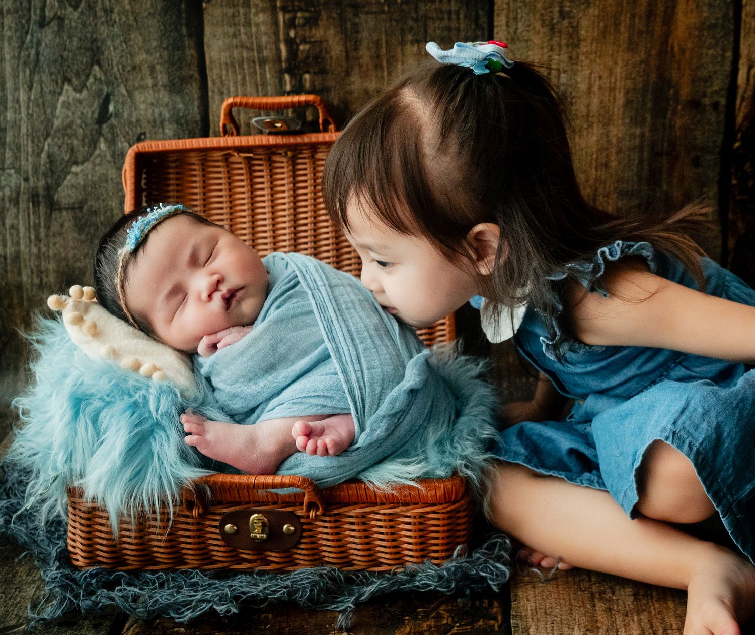 A young girl in a blue dress kisses a sleeping newborn baby wrapped in teal fabric inside a wicker suitcase.