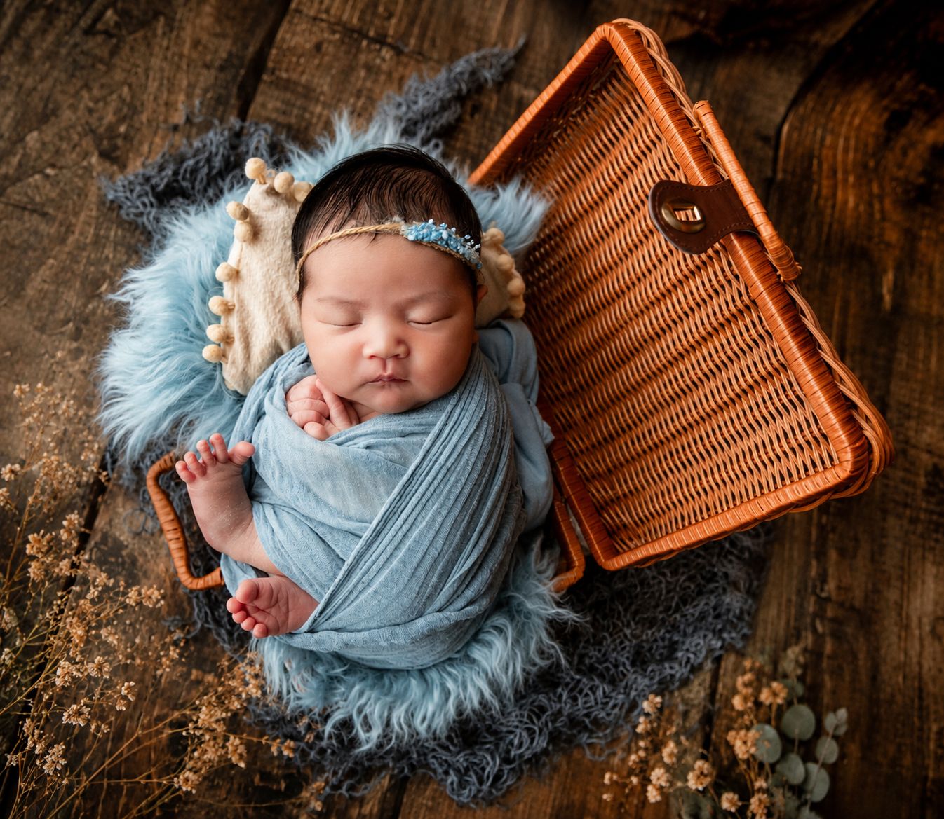 Newborn baby swaddled in blue wrap sleeping peacefully in a wicker basket on a rustic wooden background.