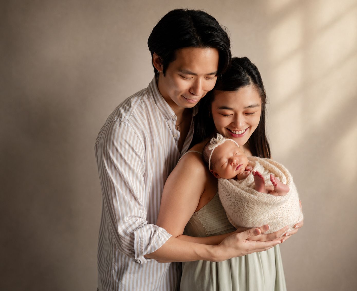 Happy Asian couple smiling at their newborn baby wrapped in a cream blanket during a studio portrait session.