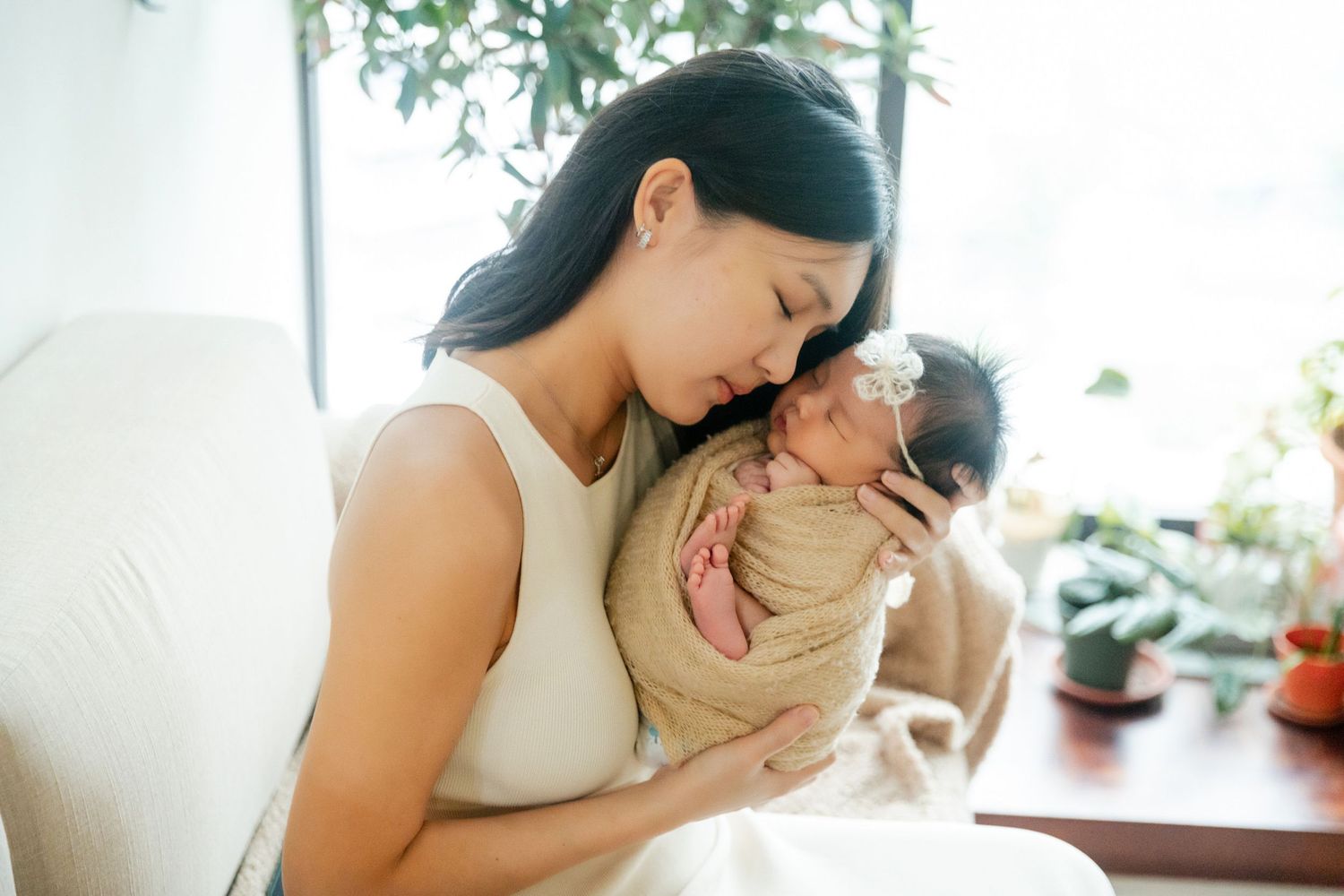 A mother tenderly holds her swaddled newborn baby close, sharing a peaceful moment in a bright, plant-filled room.