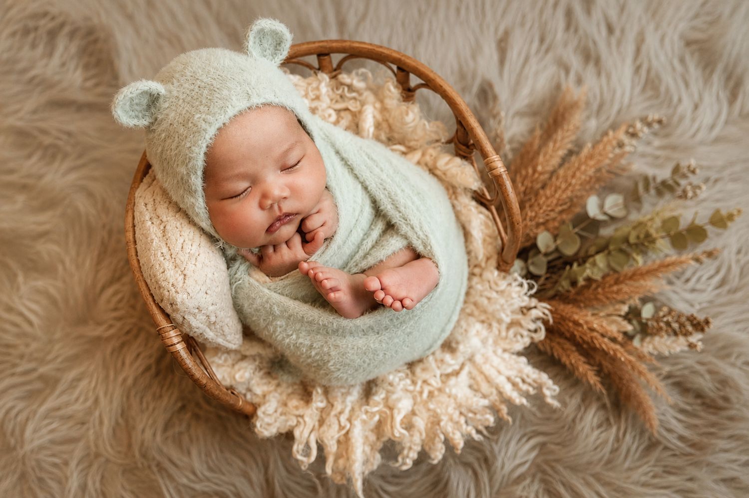 Newborn baby sleeping peacefully in a wicker basket, wrapped in mint green knit, wearing a bear bonnet.