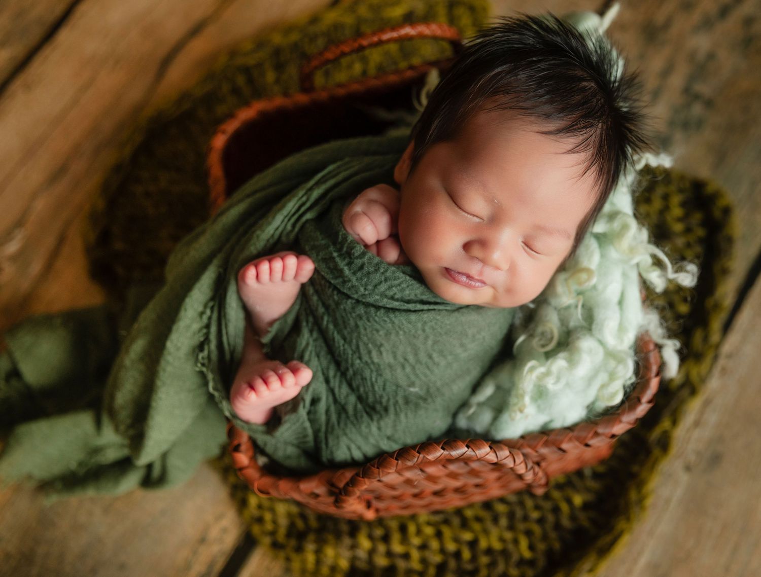 Newborn baby sleeping peacefully in a wicker basket wrapped in a green swaddle blanket.