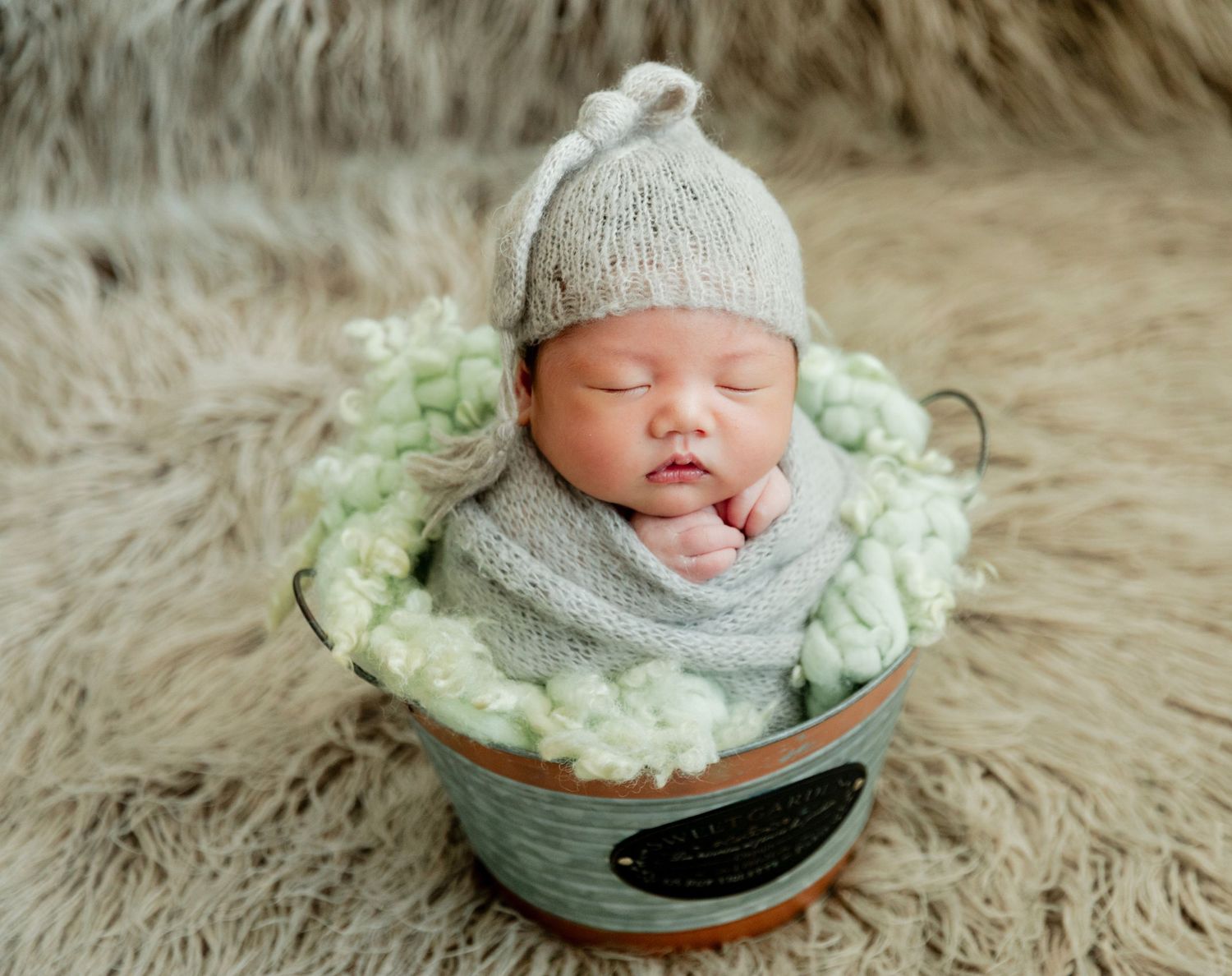 Sleeping newborn baby wrapped in gray knit swaddle and hat, nestled in a rustic bucket with green floral accents.