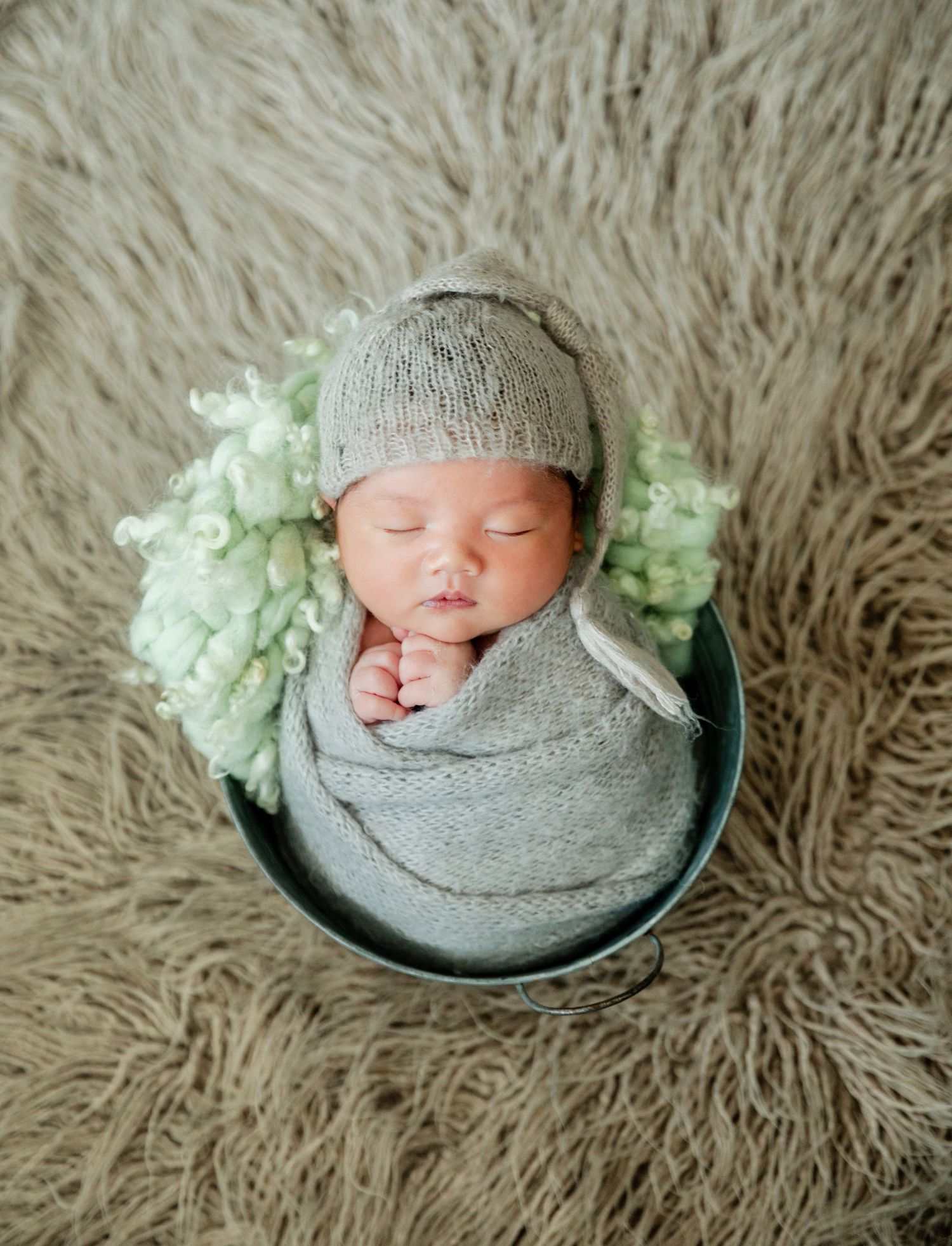 Sleeping newborn baby swaddled in gray wrap and knit hat, nestled in a bucket with green floral accents.