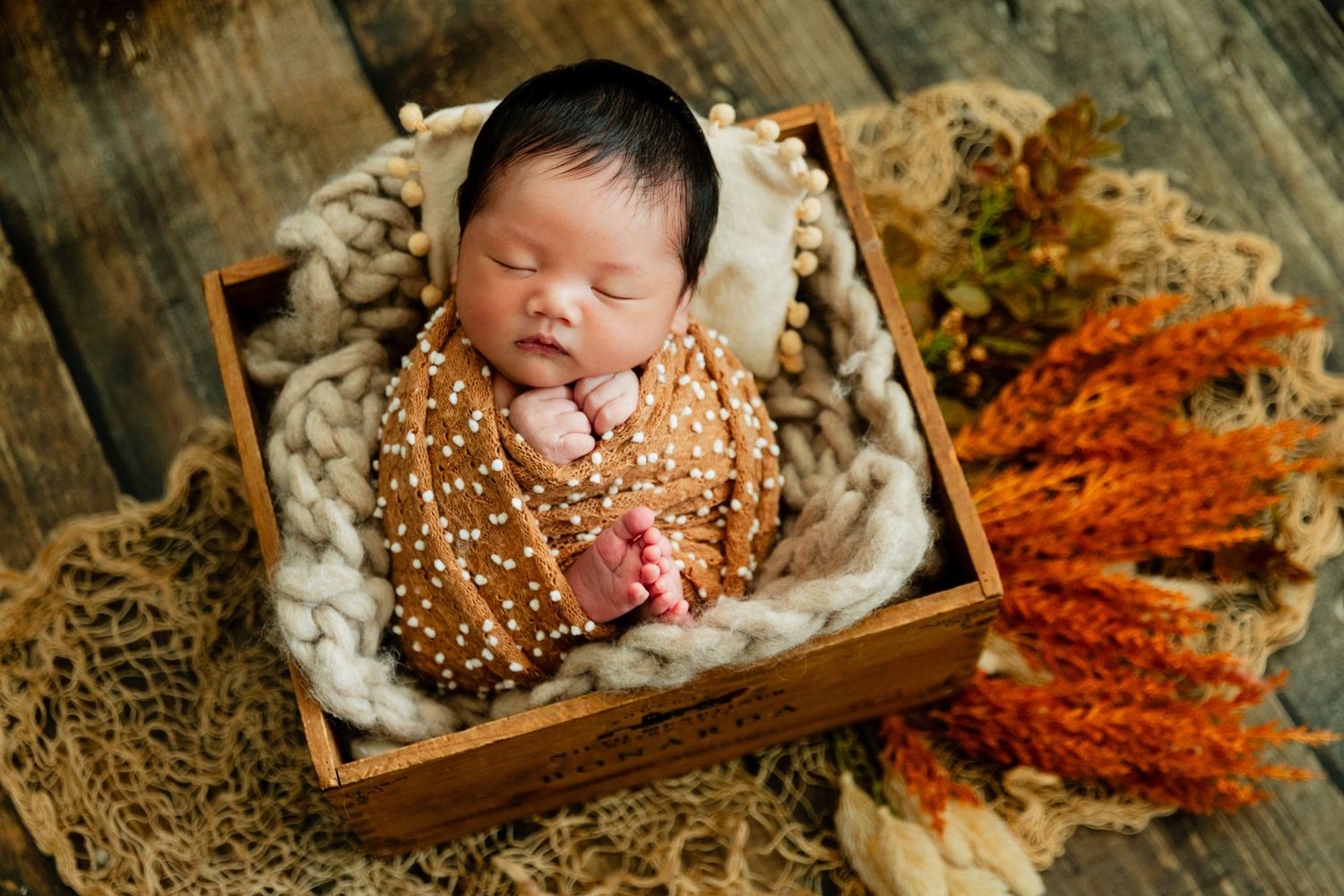 Sleeping newborn wrapped in amber swaddle inside a rustic wooden box with autumn florals.