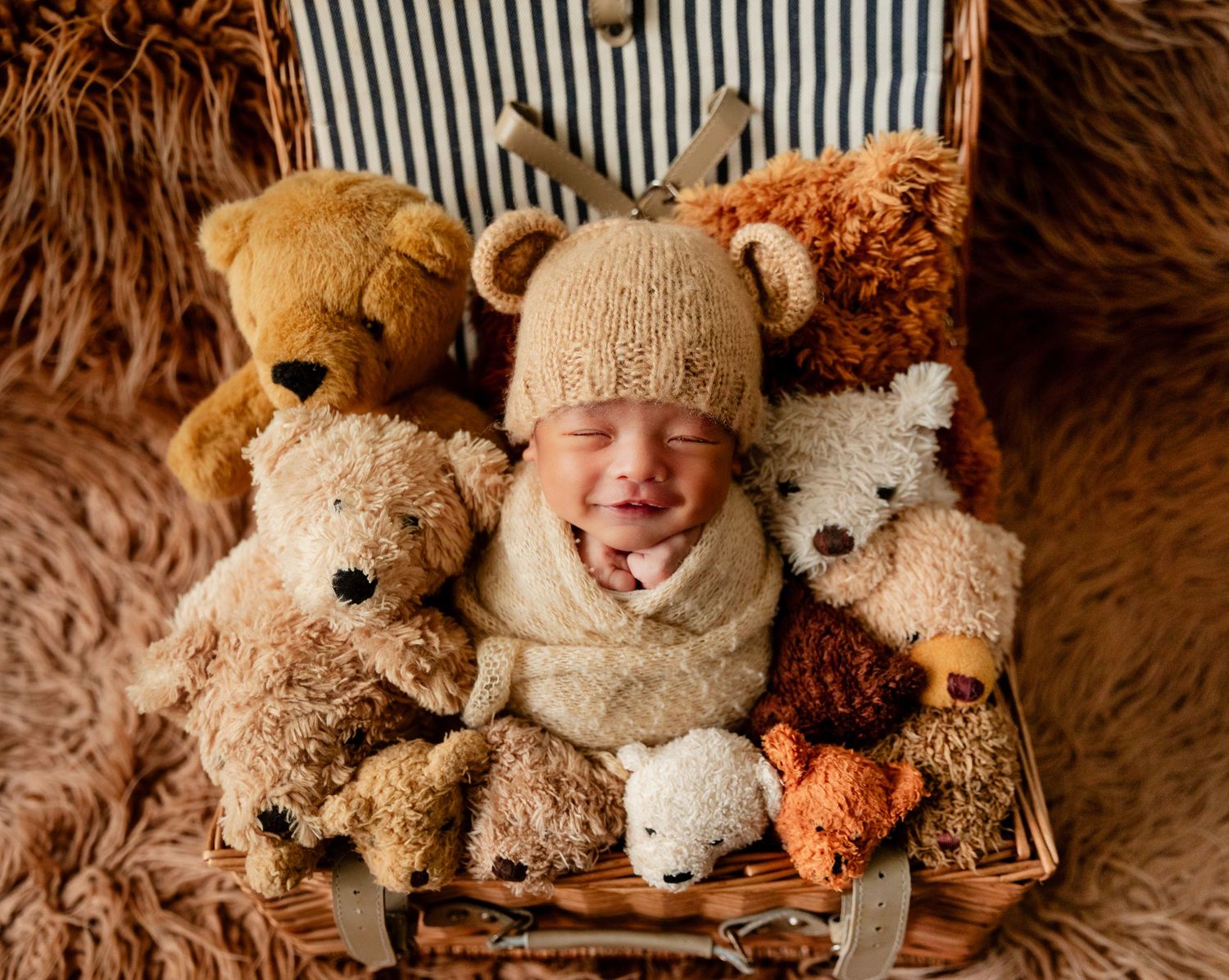 Smiling newborn baby in bear hat surrounded by teddy bears in a wicker basket.