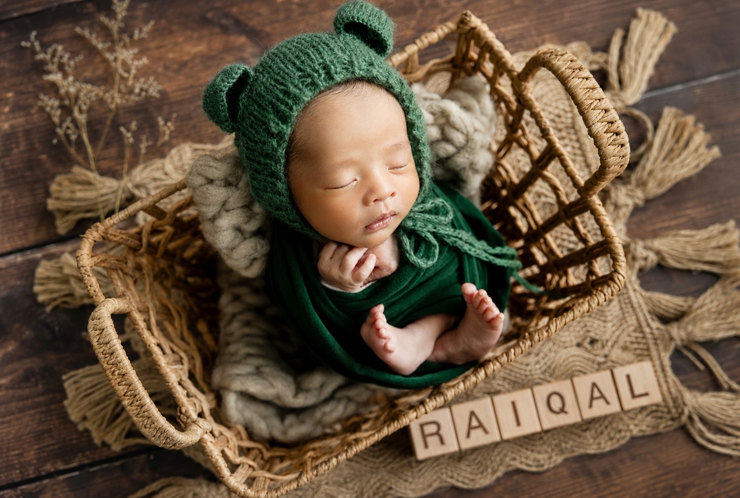 Newborn baby named Raiqal sleeping in a wicker basket wearing a green knit bear bonnet with name blocks.