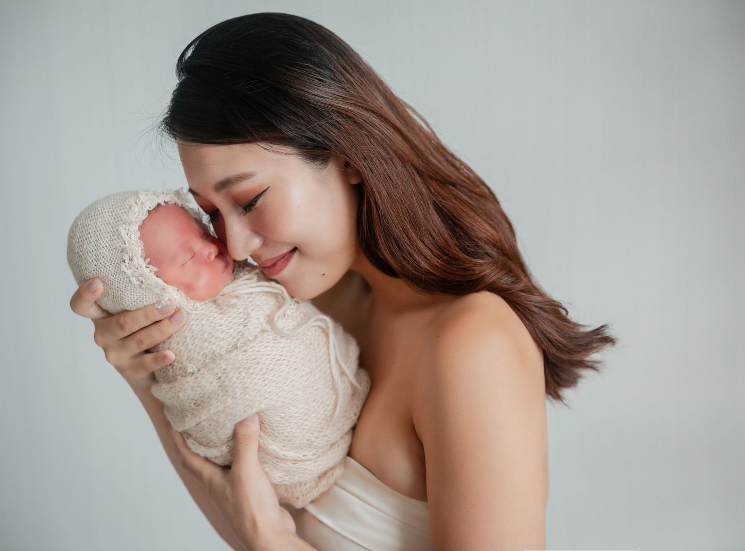 A young Asian woman tenderly kisses a sleeping newborn baby wrapped in a soft beige knit blanket.