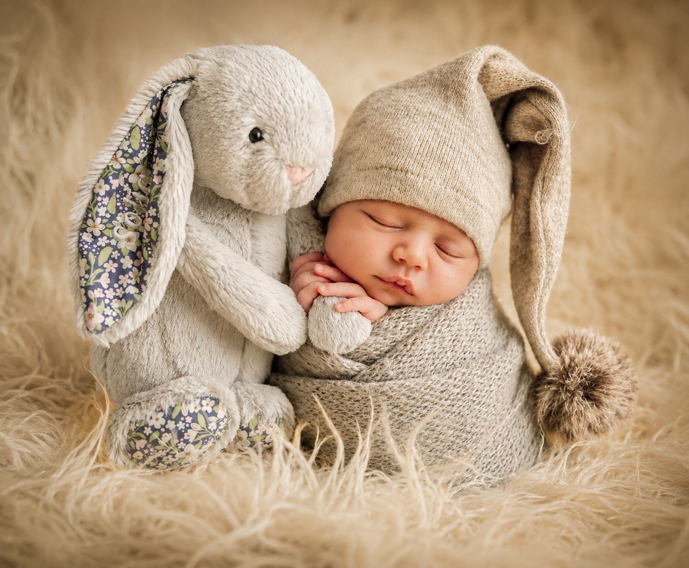 Sleeping newborn baby in bunny hat snuggled with stuffed rabbit toy on soft cream fur blanket.