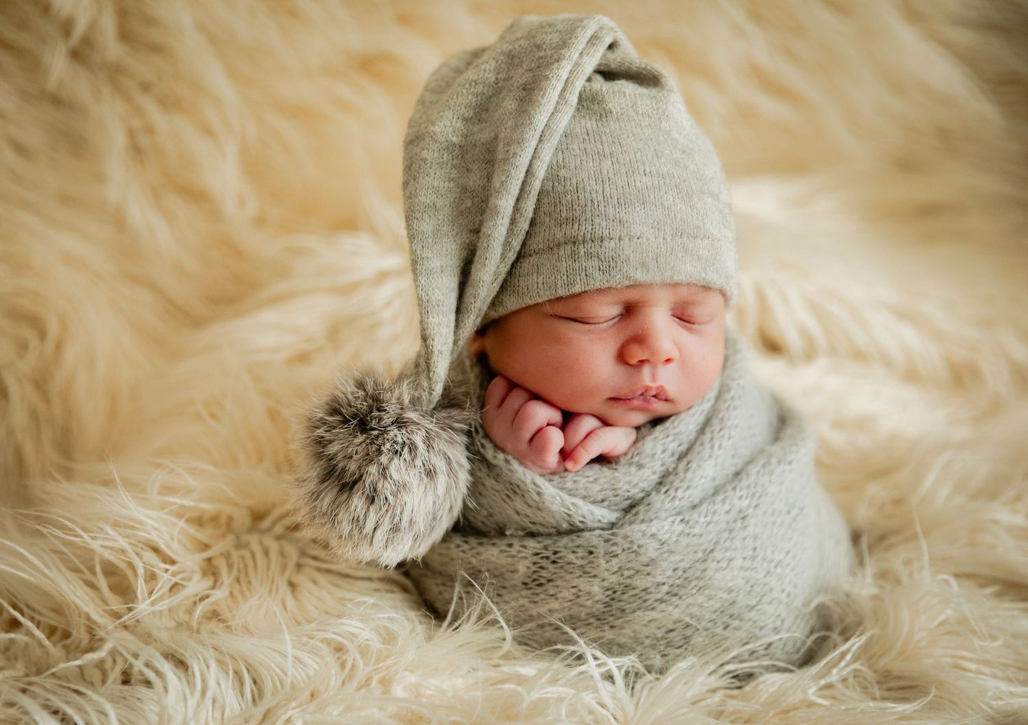Newborn baby sleeping peacefully wrapped in gray knit blanket wearing a cute pointy hat with pom-pom on fluffy fur rug.