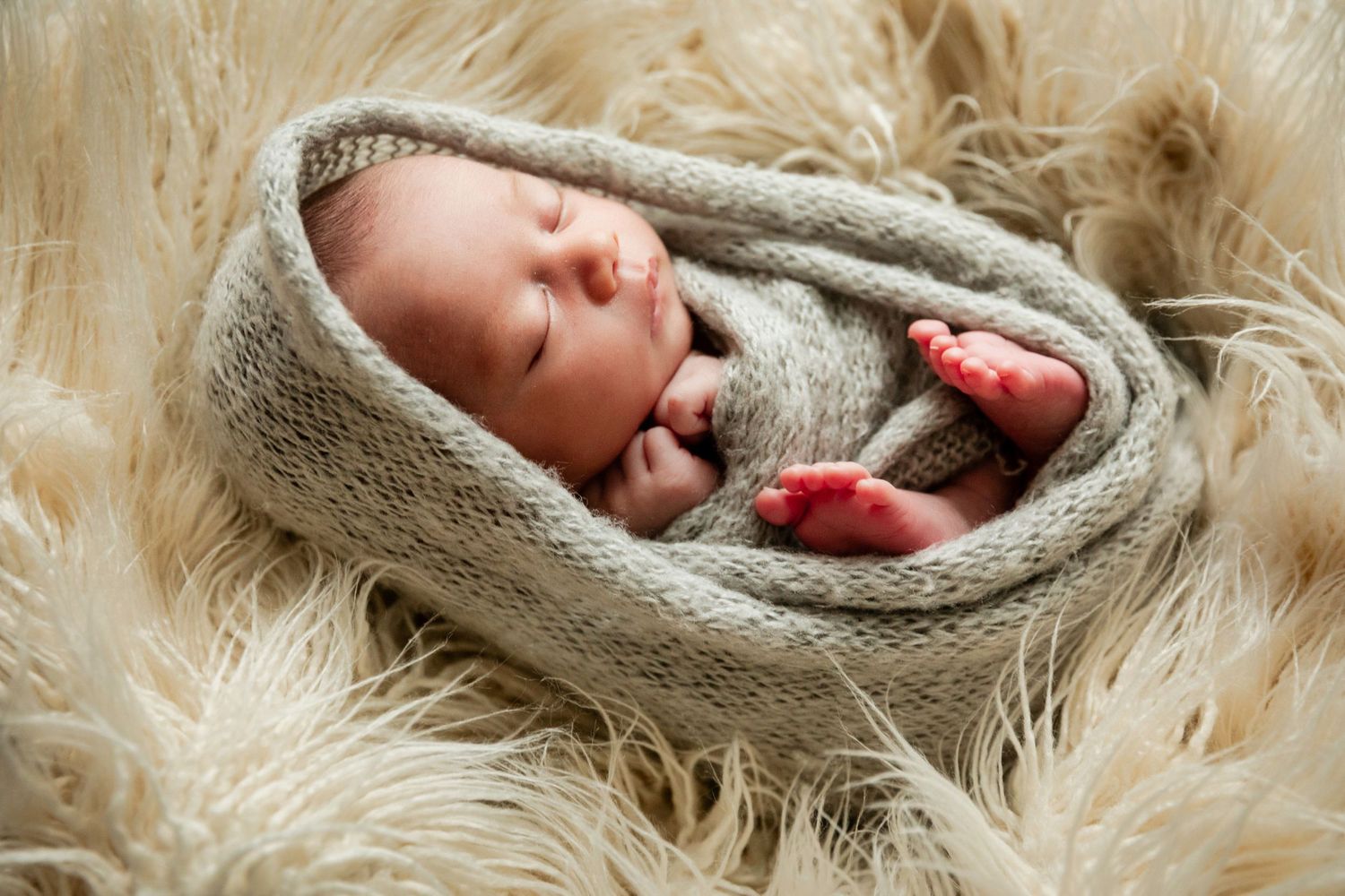 Peaceful newborn baby swaddled in a soft gray knit wrap, sleeping on fluffy cream fur, with tiny pink toes peeking out.