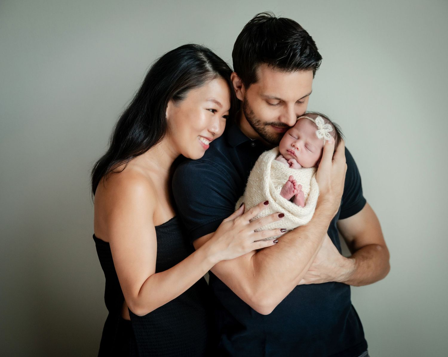A couple in black outfits smiling and holding their swaddled newborn baby during a professional studio portrait session.
