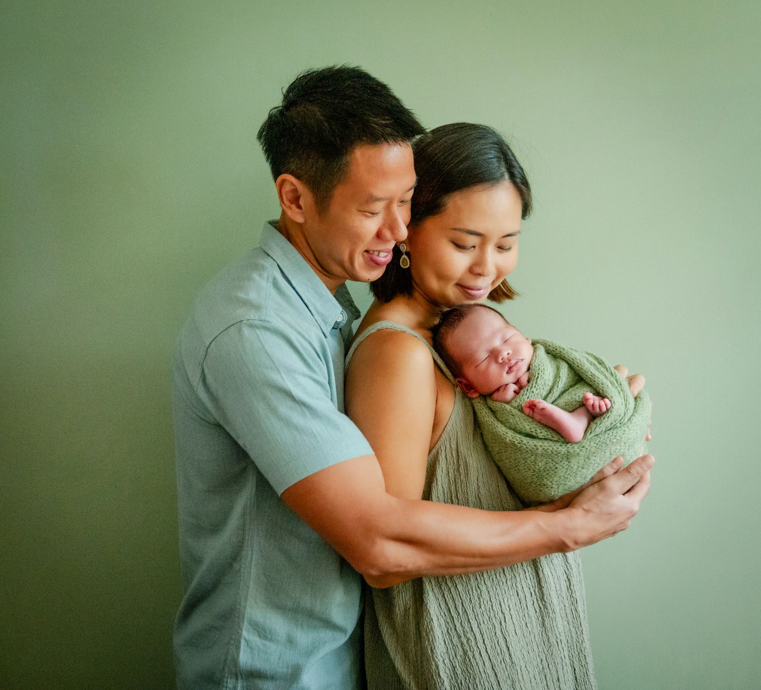 Happy parents in green outfits hold their swaddled newborn baby against a sage green studio backdrop.