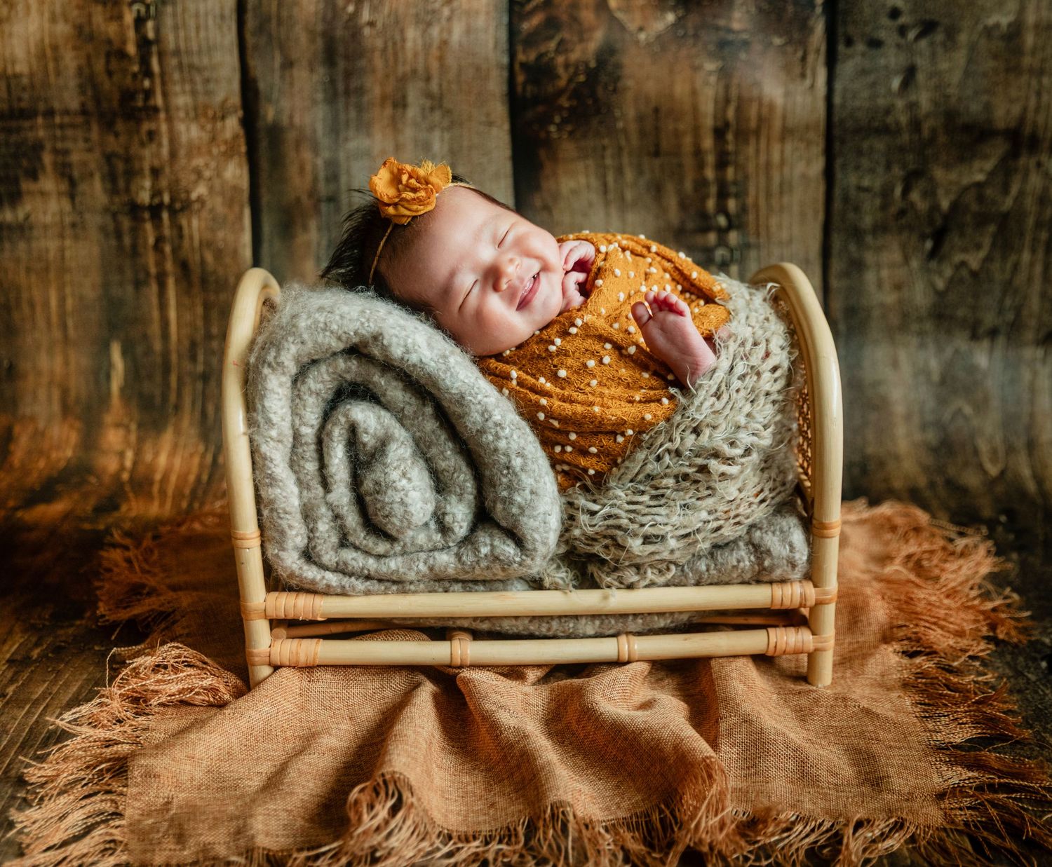 Smiling newborn baby in orange outfit sleeping peacefully in a small rattan bed with cozy blankets.