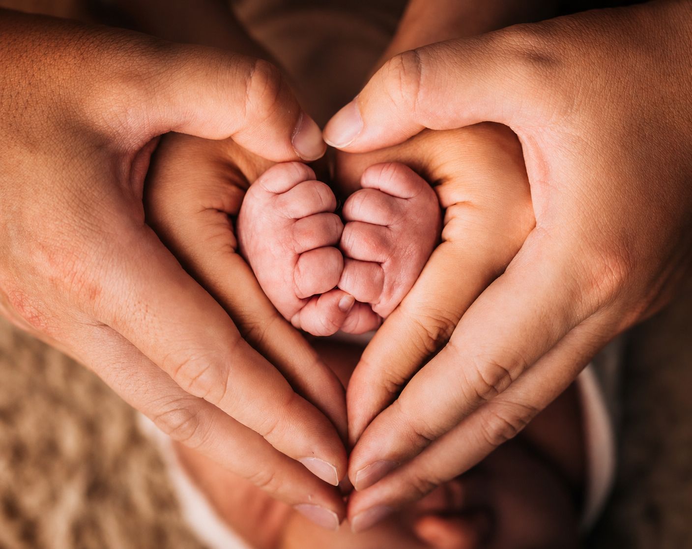 Adult hands forming a heart shape cradling tiny newborn baby feet in a tender, loving gesture.
