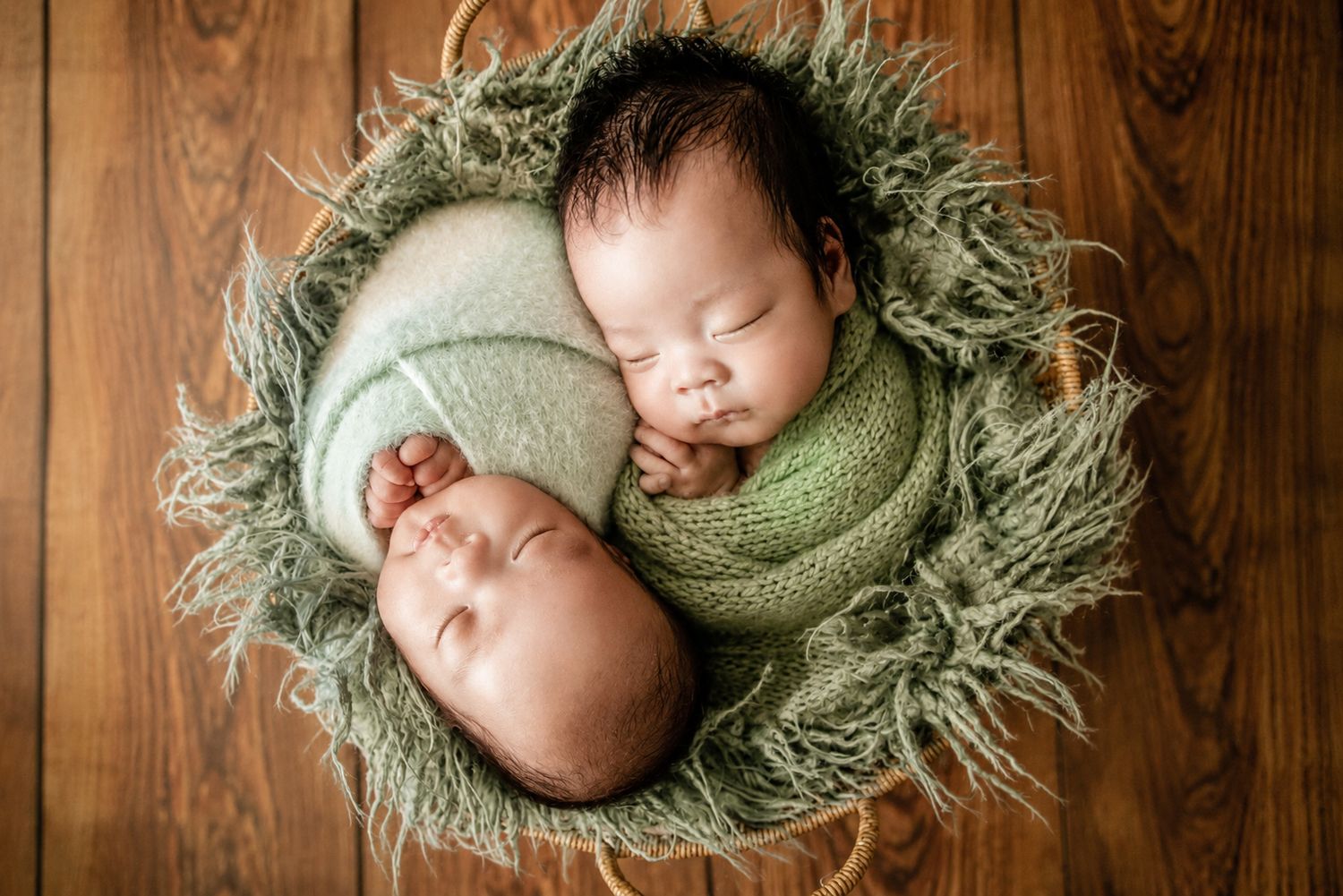 Two newborn twins swaddled in green wraps sleeping peacefully in a cozy basket on a wooden floor.