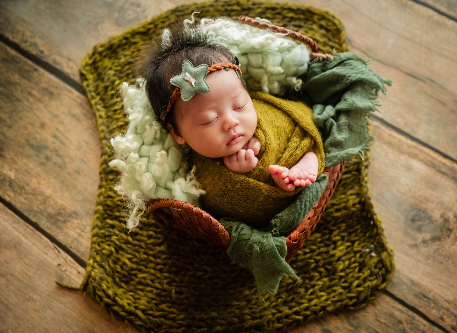 Newborn baby girl sleeping peacefully in a green knit basket with floral headband and cozy wrap.