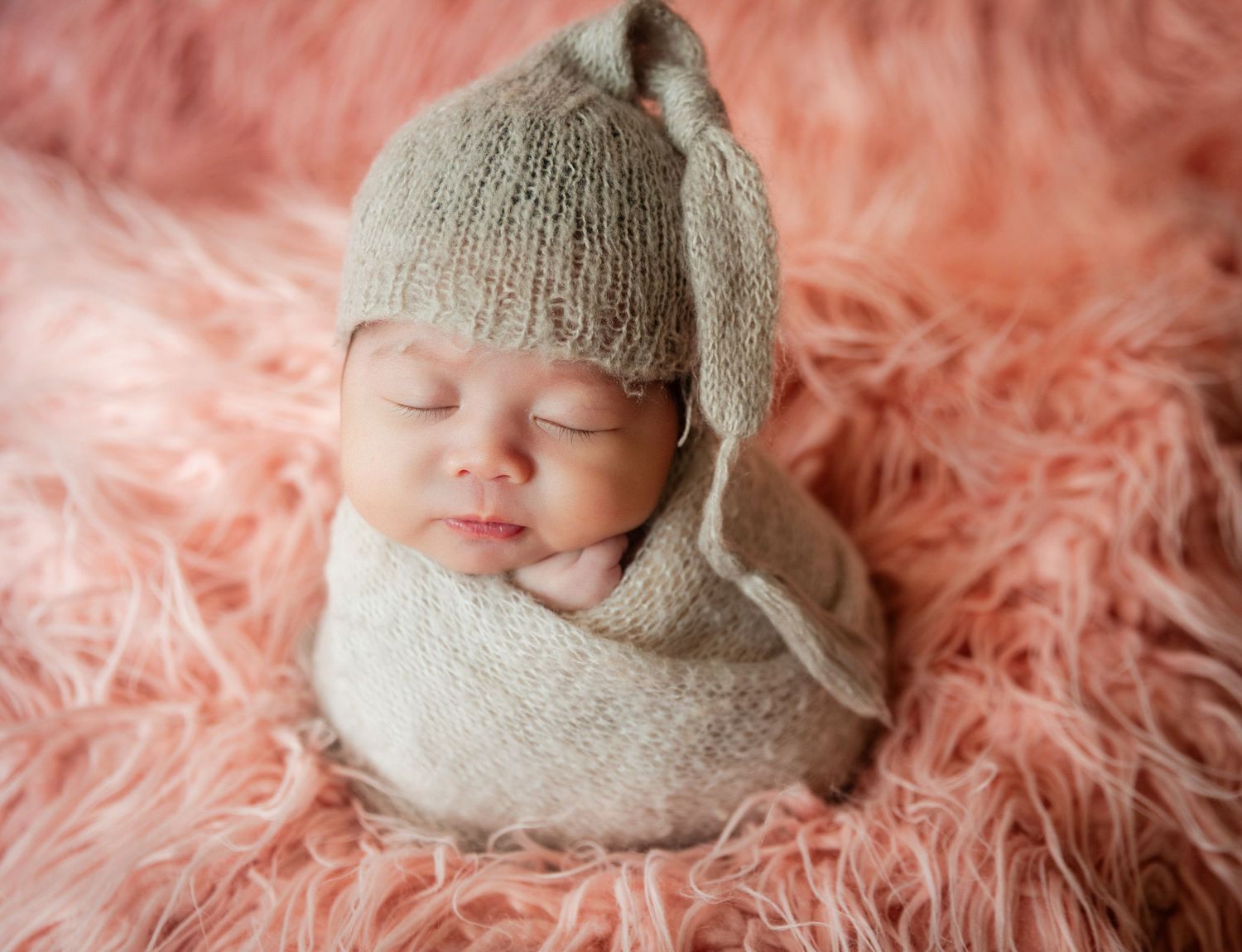 Sleeping newborn baby wrapped in a soft beige knit swaddle and hat, resting on a fluffy pink fur blanket.