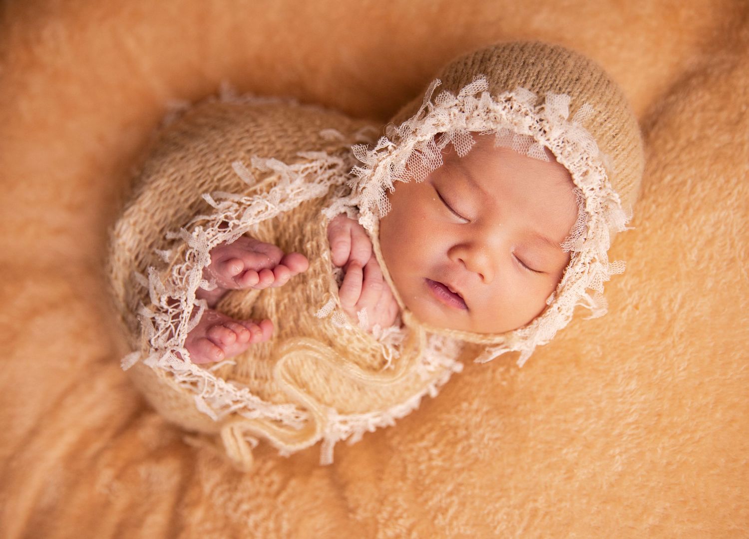 Sleeping newborn baby wrapped in cream knit swaddle and lace bonnet on soft fur blanket.
