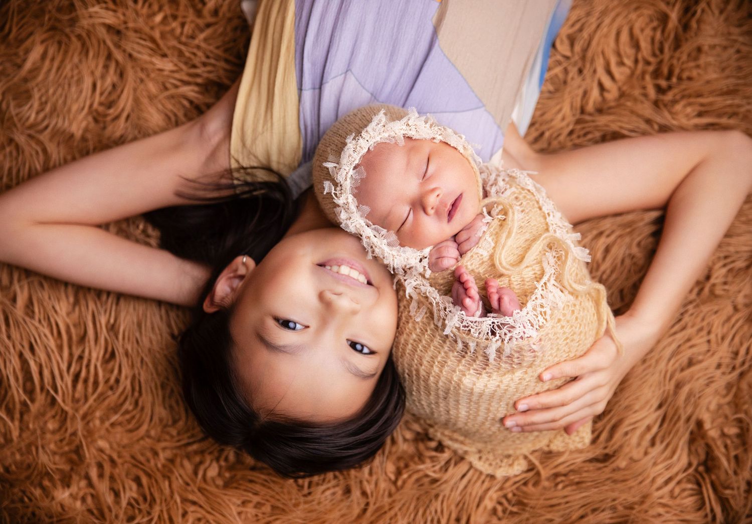 Mother lying on brown fur rug smiling beside her swaddled newborn baby wrapped in cream knit blanket.