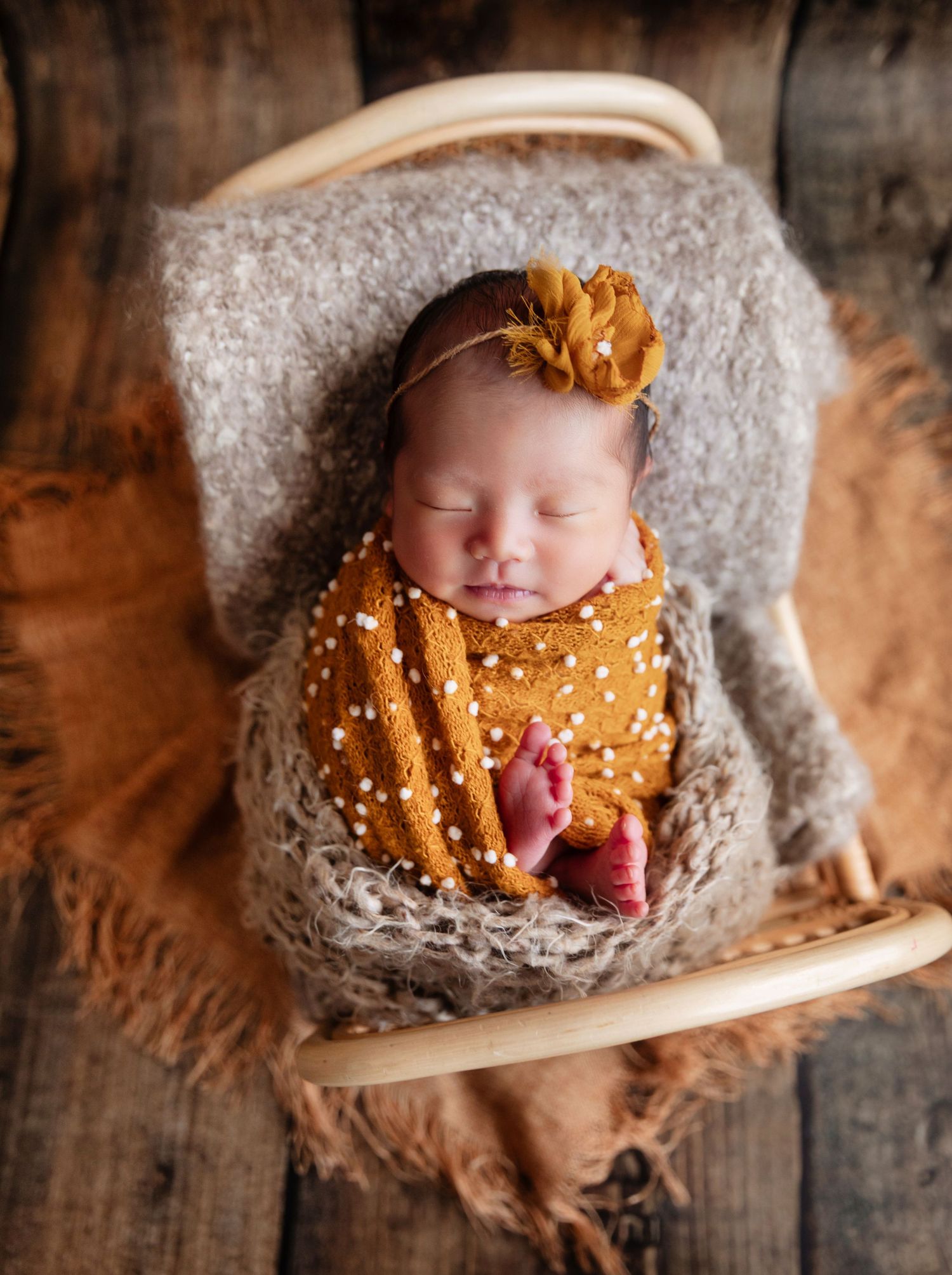 Sleeping newborn baby wrapped in mustard yellow swaddle with floral headband in a wooden bowl on rustic wood.