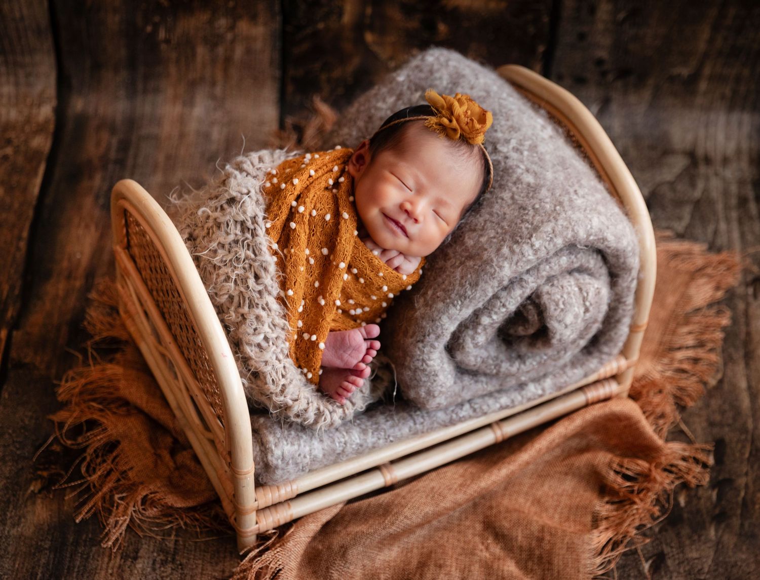 Newborn baby sleeping peacefully in a tiny wooden bed wrapped in a mustard yellow knit blanket with a floral headband.