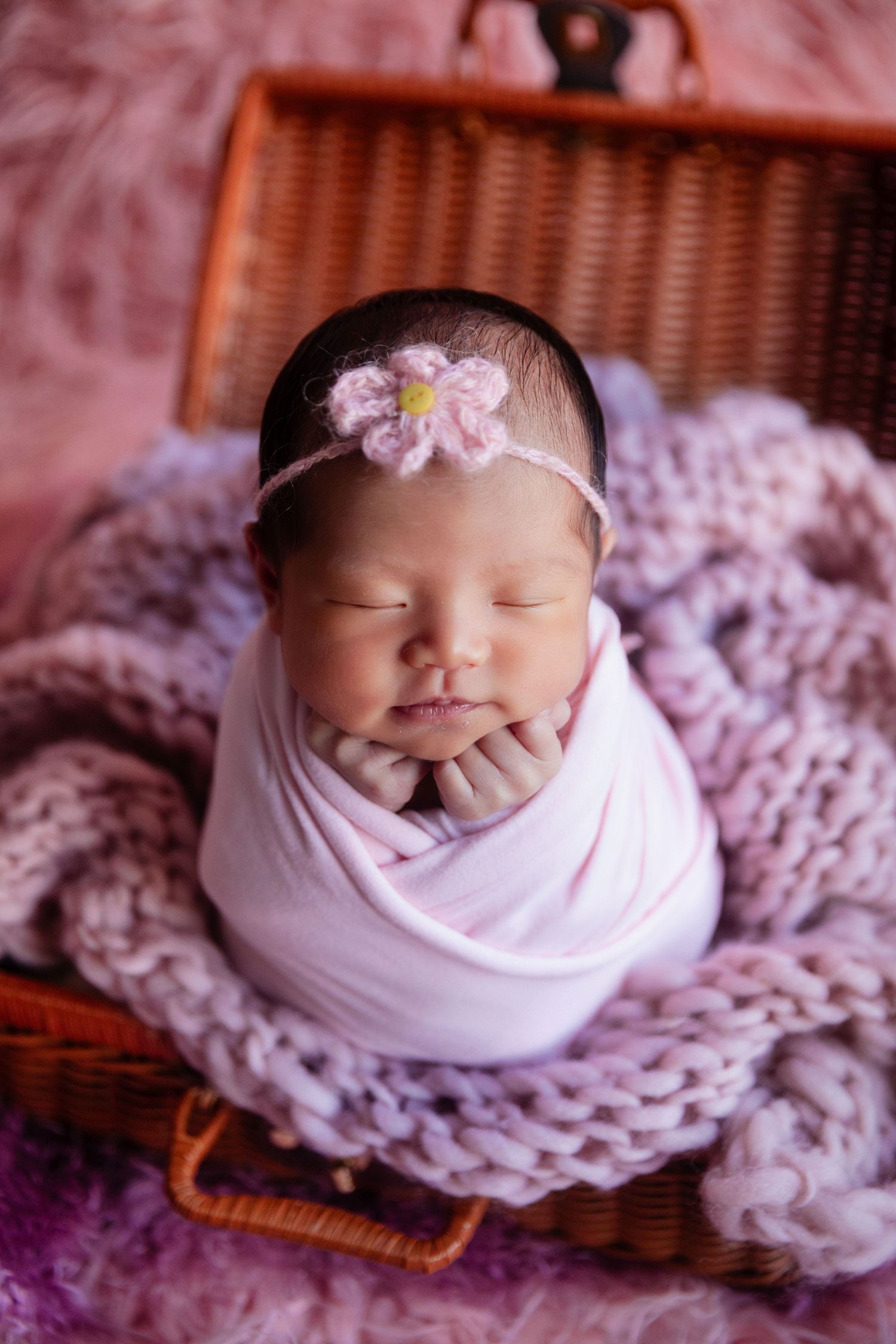 Sleeping newborn baby girl swaddled in pink wrap with floral headband, resting in wicker basket with knit blanket.