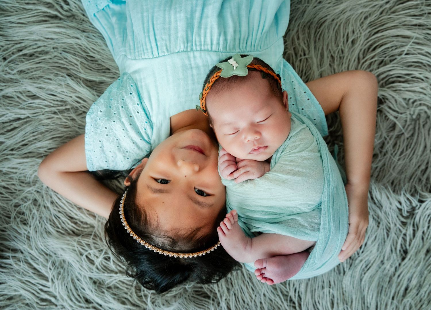 A mother and newborn baby in matching teal outfits lying on a gray fluffy rug in a newborn photography session.