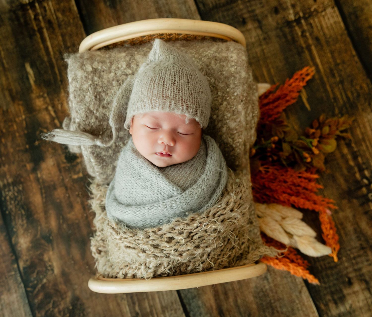 Sleeping newborn baby wrapped in gray knit swaddle wearing a knit hat, nestled in a wooden mini bed.