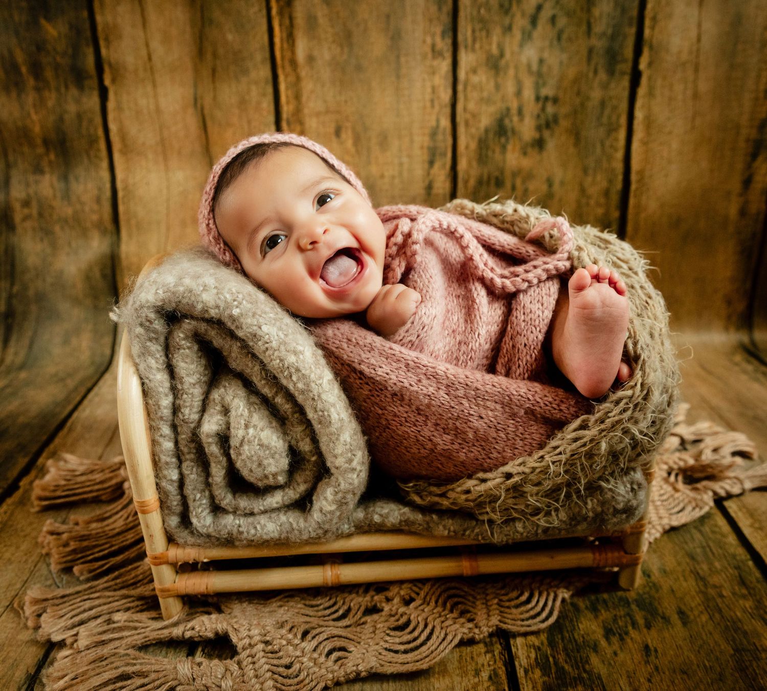 Smiling newborn baby wrapped in pink knit blanket lying in a rustic wooden crate with soft gray blankets.
