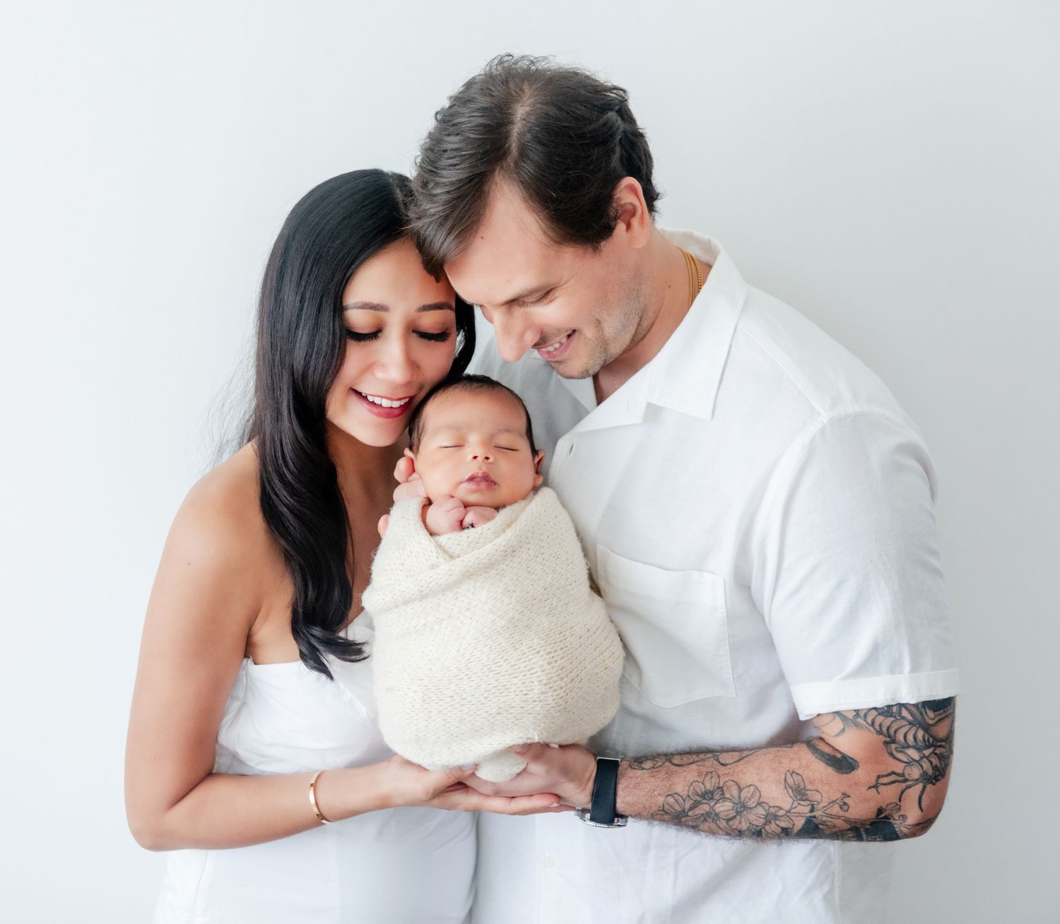 Happy new parents smiling and holding their swaddled newborn baby against a white background.