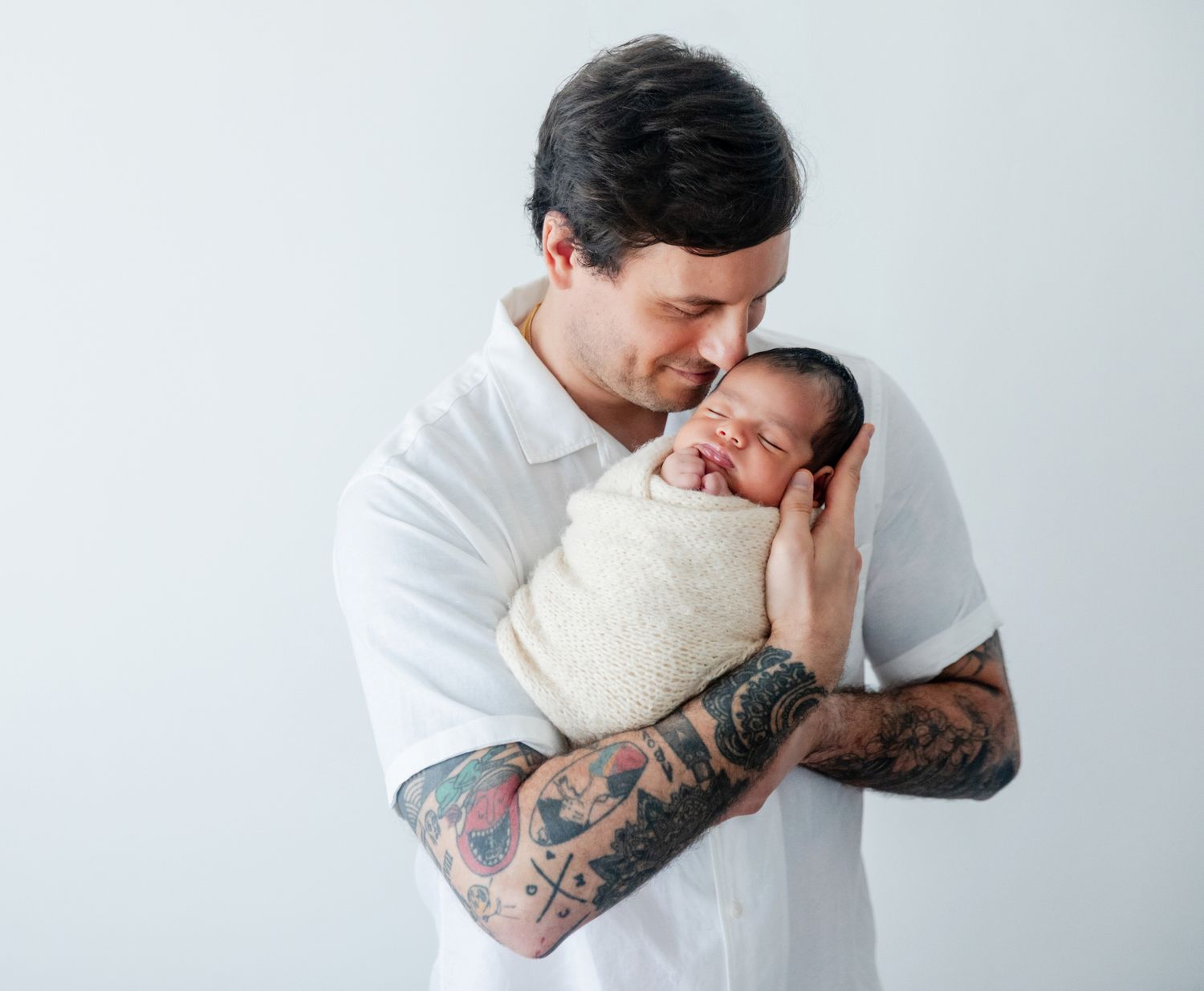 Tattooed father in white shirt tenderly holding sleeping newborn baby wrapped in white swaddle.