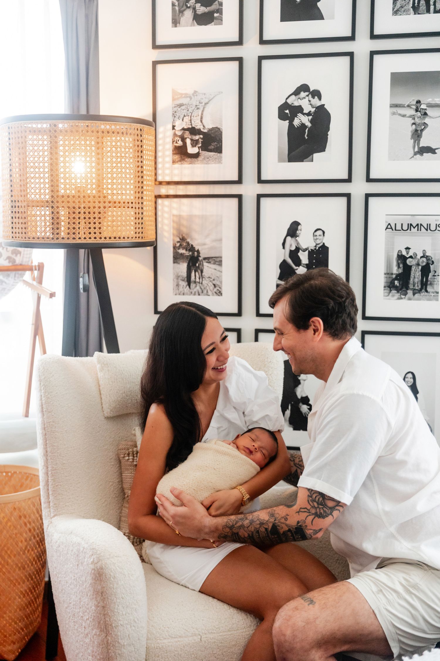 New parents admire their swaddled newborn in a white nursery chair surrounded by black and white family photos.