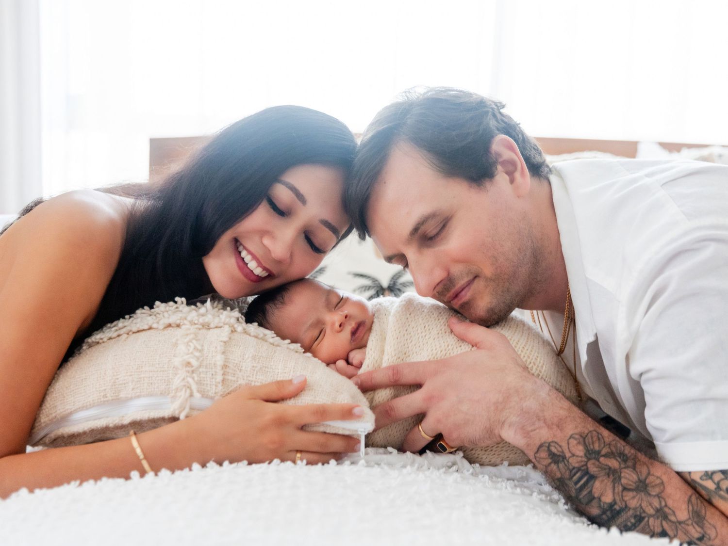 New parents lovingly gaze at their sleeping newborn baby on a white bed in a bright, cozy room.