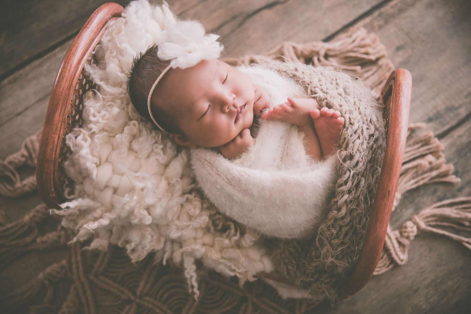Newborn baby sleeping peacefully in a wooden basket wrapped in white, adorned with a floral headband.