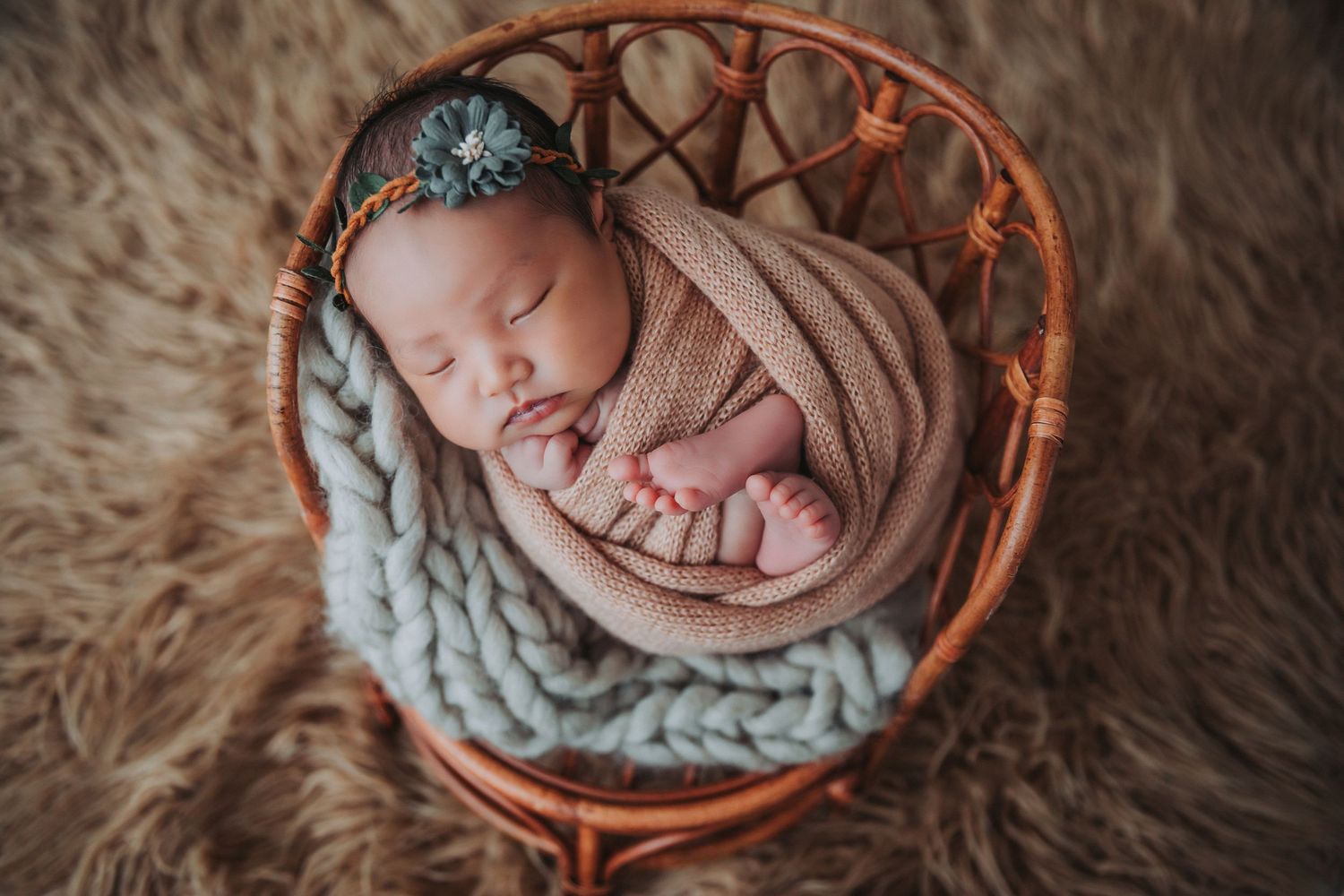 Sleeping newborn wrapped in beige swaddle nestled in a wicker basket with gray knit blanket.
