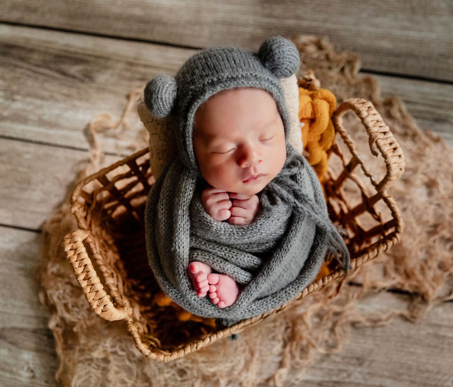 Newborn baby sleeping peacefully in a wicker basket, wrapped in a gray knit blanket and bear bonnet hat.